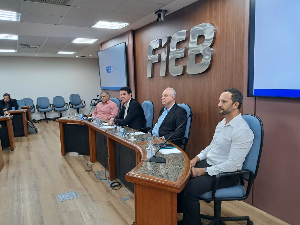 Imagem de quatro homens presentes em uma mesa, com a parede ao fundo mostrando a palavra FIEB, além de duas telas.