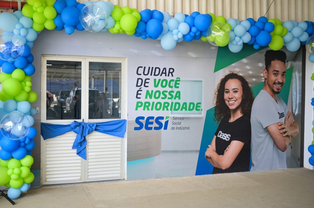 Entrada de uma unidade de programa do SESI de Atenção Primária à Saúde. A porta está envolta por um laço azul, além de bolas nas cores azul escuro, verde cana e azul claro, indicando que é uma inauguração.
Na parede, um painel dizendo "Cuidar de você é nossa prioridade", embaixo a marca do SESI.